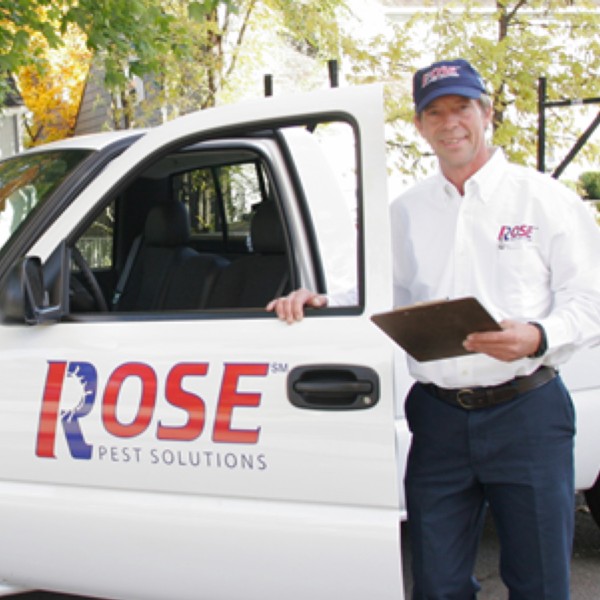 A person wearing a "Rose Pest Solutions" uniform stands by a company truck holding a clipboard.