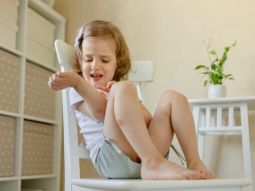 A young child sits on a white chair indoors, looking at and touching a red mark on her arm, perhaps wondering why are mosquitos attracted to me, with a small white table and potted plant in the background.