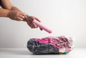 A person uses a pink manual air pump to inflate a rolled, multicolored vacuum-sealed bag on a white surface, perhaps storing items while wondering, do mothballs work to keep bugs away?.