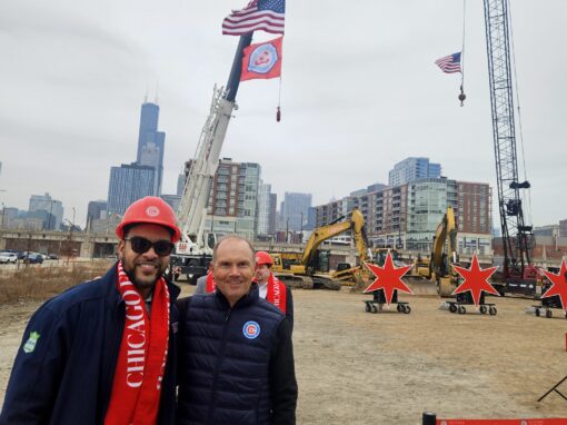 Wesley and Joe in Chicago Fire FC gear stand at Chicago Fire construction site with cranes, an American flag, a Chicago Fire FC flag, and red star displays in the background. City skyline visible.