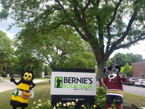 Two mascots, a bee and a wolf in a hockey jersey, stand beside a sign for Bernie’s Book Bank under a tree on a grassy area with flowers, celebrating their Chicagoland literacy partnership.