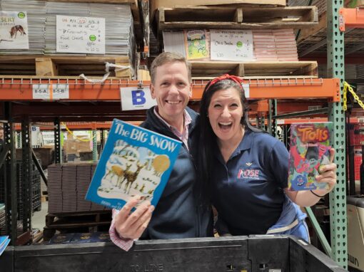 Two people stand in a warehouse, smiling and holding up books titled 