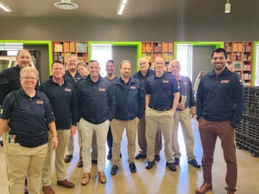 A group of twelve people wearing matching navy shirts and khaki pants pose together and smile in a workplace setting, representing the Chicagoland literacy partnership, with shelves and bins in the background.