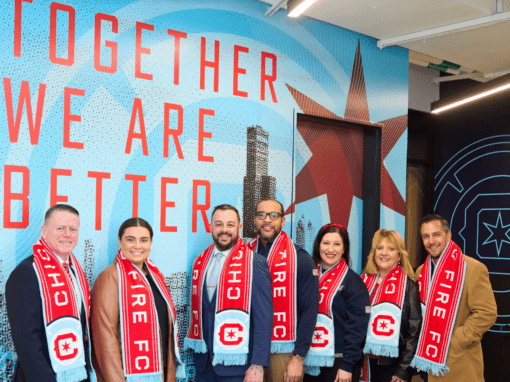 Rose and Fire team in Chicago Fire FC scarves stand in front of a wall with the text “Together We Are Better” and Chicago-themed graphics, celebrating the spirit of community and the strength of their Chicago Fire FC partnership.