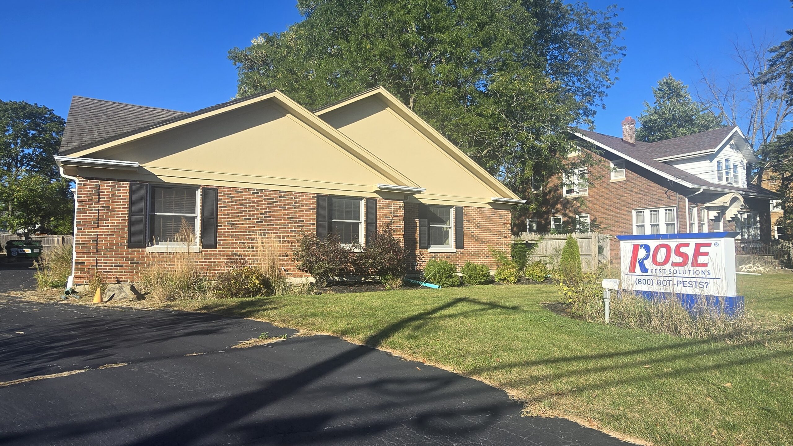 A brick house with a tan roof sits beside a driveway. A "Rose Pest Solutions" sign is posted on the lawn. Trees and another house are visible in the background.