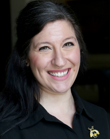 A woman with long dark hair and a black top smiles at the camera, proudly wearing a bee-shaped brooch; she's known as the Chicago Bug Girl.