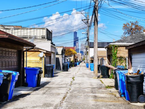 A city alley in Chicago lined with blue and black garbage bins, garages on both sides, and overhead utility wires, with a tall skyscraper visible in the distance under a blue sky—a possible site for rat birth control initiatives.